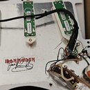 Fotografie zachycuje zadní stranu bílého trupu elektrické kytary s odhalenou elektronikou a stříbrnou stínící vrstvou. Jsou zde tři snímače s nápisy…