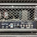 Detail zadní strany kytarového komba s černým texturovaným potahem a ochrannou kovovou mřížkou, za kterou jsou vidět elektronické součástky včetně va…