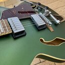 Detailní pohled na tělo zelené elektrické kytary s matným povrchem a vyřezaným otvorem (semi-hollow). Nástroj má dva chromované snímače s krytkami, h…