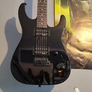 Černá elektrická kytara s lesklým povrchem a hnědým hmatníkem. Nástroj má dva humbuckery, jeden ovládací knoflík hlasitosti a jeden přepínač. Na těle…