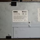 Fotografie zachycuje spodní stranu bílého elektronického zařízení. Uprostřed je nalepený štítek se značkou KORG, označením MODEL: AX5G a sériovým čís…