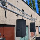 Dva černé reproduktory s kovovými mřížkami jsou zavěšeny na hliníkovém nosném trussu. Reproduktory mají na bocích úchyty a jsou upevněny pomocí černý…