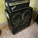 Černý kytarový zesilovač s hlavou umístěnou na basovém boxu. Na ovládacím panelu hlavy je nápis Fender a označení 1000. Box obsahuje čtyři reprodukto…