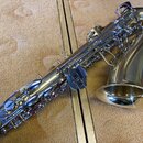Zlatý saxofon s kovovým povrchem, položený na béžovém podkladu. Nástroj má stříbrné klapy a mechanismus, na kterém nejsou vidět žádné značky ani nápi…