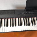 Černý digitální klavír s bílými a černými klávesami. Na horním ovládacím panelu jsou nápisy MASTER VOLUME, DEMO/SONG, SELECT, METRONOME, PIANIST RHYT…