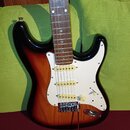 Elektrická kytara typu Stratocaster s korpusem v barevném přechodu od černé přes červenou k žluté. Nástroj má bílý pickguard, tři krémové snímače a h…