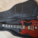 Červená elektrická kytara s černým pickguardem v černém polstrovatém obalu. Na hlavě je nápis "J. S. BACHMAN". Tělo kytary má dva humbuckery, čtyři o…