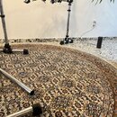 Fotografie zachycuje spodní část chromovaných stojanů na hudební nástroje umístěných na kruhovém vzorovaném koberci. Jsou vidět kovové nohy s černými…