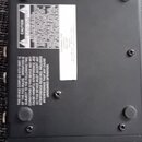 Fotografie zachycuje spodní stranu černého elektronického zařízení s matným povrchem. Na spodní straně jsou čtyři gumové nožičky a několik viditelnýc…