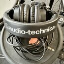 Černá uzavřená sluchátka s polstrováním z umělé kůže. Na hlavovém oblouku je bílý nápis audio-technica s logem značky. Pod sluchatky je smotaný černý…