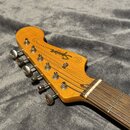 Fotografie zachycuje hlavu elektrické kytary z přírodního dřeva s nápisy Squier a JAZZMASTER. Na hlavě jsou umístěny šest kovových ladiček stříbrné b…