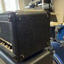 Detailní pohled na horní část kytarového komba v černém texturovaném vinylu s kovovými rohy. Na přední straně je vidět stříbrná tkaná síť a část ovlá…