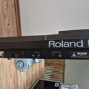 Detailní pohled na zadní panel černého elektronického nástroje. Je zde vidět bílý nápis Roland G- a štítek s označením Roland CE, specifikací napájen…