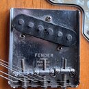 Detail kovové základní desky elektrické kytary s instalovaným snímačem. Na desce je vyražen nápis FENDER a pod ním menší text PAT. PEND. BUSHNEST 2,5…