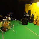 Fotografie zachycuje hudební studio s bicími nástroji a několika kytarami. V popředí je bicí sada s tmavě zeleným basovým bubnem, dvěma béžovými tomm…