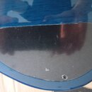 Detail zadní strany modré elektrokitary s viditnou strukturou dřeva. Na těle je přišroubovaná oválná černá plastová krytka elektroniky. Povrch krytky…