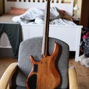 Fotografie zachycuje zadní stranu elektrické basové kytary s dřevěným korpusem a hnedým povrchem. Nástroj má čtyři struny, tmavý hmatník a na zadní s…