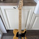 Elektrická kytara typu Telecaster v přirozené dřevěné barvě s černým pickguardem. Na hlavě hmatníku je vidět nápis „Fender“ a modelové označení „TELE…