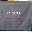 Černý textilní kryt na hudební zařízení s bílým nápisem CATALYST uprostřed. Materiál vykazuje vrásky a mírné pomačkání. Kryt je navlečen na stříbrný…