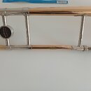 Detailní pohled na část mosazného a stříbrného posuvu z trumpety. Na stříbrném ventilovém mechanismu je černý kulatý štítek s nápisem „Arnold & Sons…