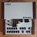 Bíle-šedý organický expander v dřevěném rámu s nápisy OB-3 Drawbar Organ Expander a logem Oberheim. Přístroj obsahuje tři sady tahových regulátorů (d…