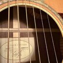 Detail pohledu do ozvukové otvory akustické kytary s viditelnými strunami a hmatníkem. Uvnitř korpusu je nalepený štítek s nápisy TAYLOR GUITARS, MOD…