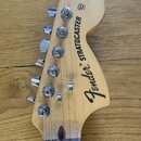 Hlava elektrické kytary z přírodního světlého dřeva s chromovanými ladičkami. Na povrchu je černý nápis Fender® STRATOCASTER a menší text ORIGINAL Co…