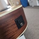 Detailní záběr na zadní část akustické kytary s hnědým dřevěným povrchem a bílým lemováním. V těle je osazen černý plastový ovládací panel s jackovým…