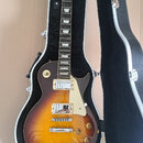 Elektrická kytara typu Les Paul s barevným přechodem od žluté k tmavě hnědé v hardcase s černým vnitřkem. Na hlavě je nápis "Vantage". Kytara má dva…