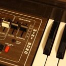 Detailní pohled na ovládací panel a klávesy elektronického klavíru s hnědým dřevěným povrchem. Na panelu jsou nápisy ACCOMPANIMENT, DRUMS, MINI BAND,…