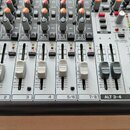 Detailní pohled na horní panel míchacího pultu stříbrné barvy. Jsou vidět posuvníky hlasitosti, otočné knoflíky a tlačítka s nápisy MUTE, CLIP a SOLO…