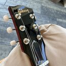 Fotografie zachycuje hlavu kytary v černé barvě s lesklým povrchem. Na horní části je zlatý nápis Gibson. Nástroj má šest ladicích kol s bílými plast…