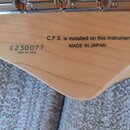 Detail dřevěného krku hudebního nástroje s viditelnou masivním kovovým hardwarem v horní části. Na dřevě jsou černé nápisy „C.F.S. is installed on th…