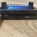 Černý elektronický přístroj s displejem a ovládacími knoflíky. Na zadní straně jsou nápisy POWER, DEVICE, CLOCK IN OUT, MIDI IN OUT, AUDIO IN 1 2 3 a…