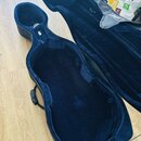Otevřený černý tvrdý kufýr na violoncello s tmavě modrou plyšovou výstělkou. Na vnitřní straně víčka je přišitý textilní štítek s bílým nápisem Bam.…