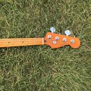 Hmat krku a hlavy basové kytary v oranžové barvě, položené na trávě. Na hlavě je nápis "Fender" a pod ním menší text "PRECISION BASS". Jsou vidět čty…