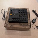 Černý sampler Digitakt značky Elektron, umístěný na hnědé kartonové krabici. Zařízení má displej, několik otočných knoflíků a mřížku bílých podsvícen…