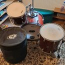 Sada bubnů v tmavě červené barvě s dřevěnou strukturou. Na basovém bubnu je nápis MAPEX a REMO, na tomaru je nápis EVANS a na druhém bubnu logo REMO.…