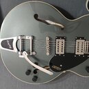 Šedo-zelená polodupatá elektrická kytara s černým pickguardem a chromovaným hardwarem. Na hrazdicím mechanismu je nápis Bigsby a na pickguardu značka…