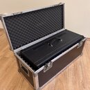 Otevřený transportní flightcase s černým povrchem a hliníkovými hranami a rohy. Uvnitř je umístěn černý kytarový kombozesilovač s rukojetí. Víko kufr…