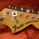 Detail hlavy elektrické kytary z přírodního dřeva. Na dřevě je černý nápis Fender STRATOCASTER a pod ním menší text PAT. 3,143,098, vpravo je nápis O…