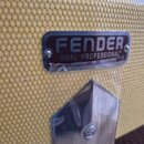 Detailní záběr na kovový štítek připevněný k žlutému texturovanému potahu zesilovače. Na štítku je černý nápis FENDER, pod ním text DUAL PROFESSIONAL…