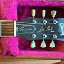Detail hlavy elektrické kytary v otevřeném kufru s růžovou plyšovou výstělkou. Na černé hlavě jsou bílé nápisy „Classic“, „Les Paul MODEL“ a vertikál…