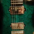 Detail elektrické kytary s tmavě zeleným povrchem s výraznou strukturou dřeva. Nástroj má dva chromované humbuckery a chromovaný kobylník. Na spodním…