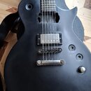 Černá elektrická kytara s matným povrchem, položená v černém obalu. Nástroj má dva humbuckery a jeden ovládací knoflík, chybí u ní plastové krytky po…