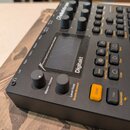 Černý digitální sampler a sekvencer s nápisem Digitakt. Zařízení má několik ovládacích knoflíků, tlačítka v různých barvách (bílá, oranžová) a malý d…