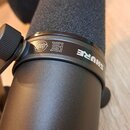 Detailní pohled na černý mikrofon s nasazenou pěnovou větrobrýkou. Na kovovém držáku je bílý nápis SHURE, značka EAC a v kruhu označení 25. Povrch mi…