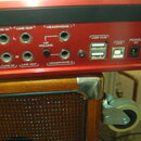 Detail zadního panelu červeného elektronického zařízení s řadou konektorů a ovládacích prvků. Jsou zde nápisy PEDAL A, MIC 1/LINE IN, LINE OUT (L, R)…
