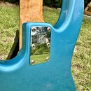 Detail zadní strany modré elektrické kytary s kovým krytem krkového spoje. Na chromovaném krytu je vyryto logo s nápisem MUSIC MAN. Povrch kytary má…