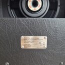 Detailní záběr na zadní část černého kytarového komba s povrchem z texturovaného vinylu. Na šasi je připevněn kovový štítek s nápisy: Model No.: G112…